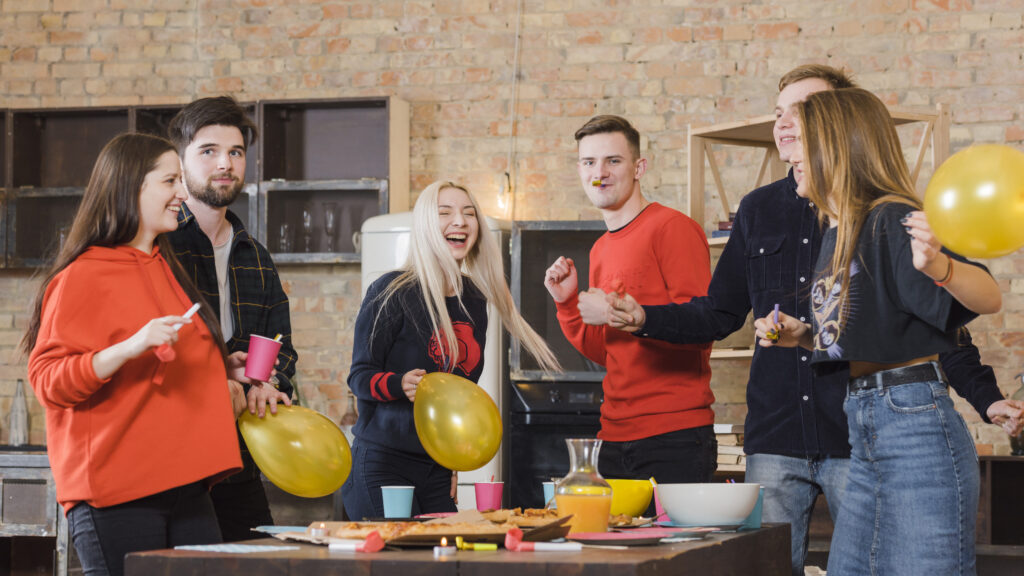 Groupe de six jeunes adultes riant et f^tant Noël avec des ballons jaunes et des boissons, ambiance conviviale d'événement intérieur.